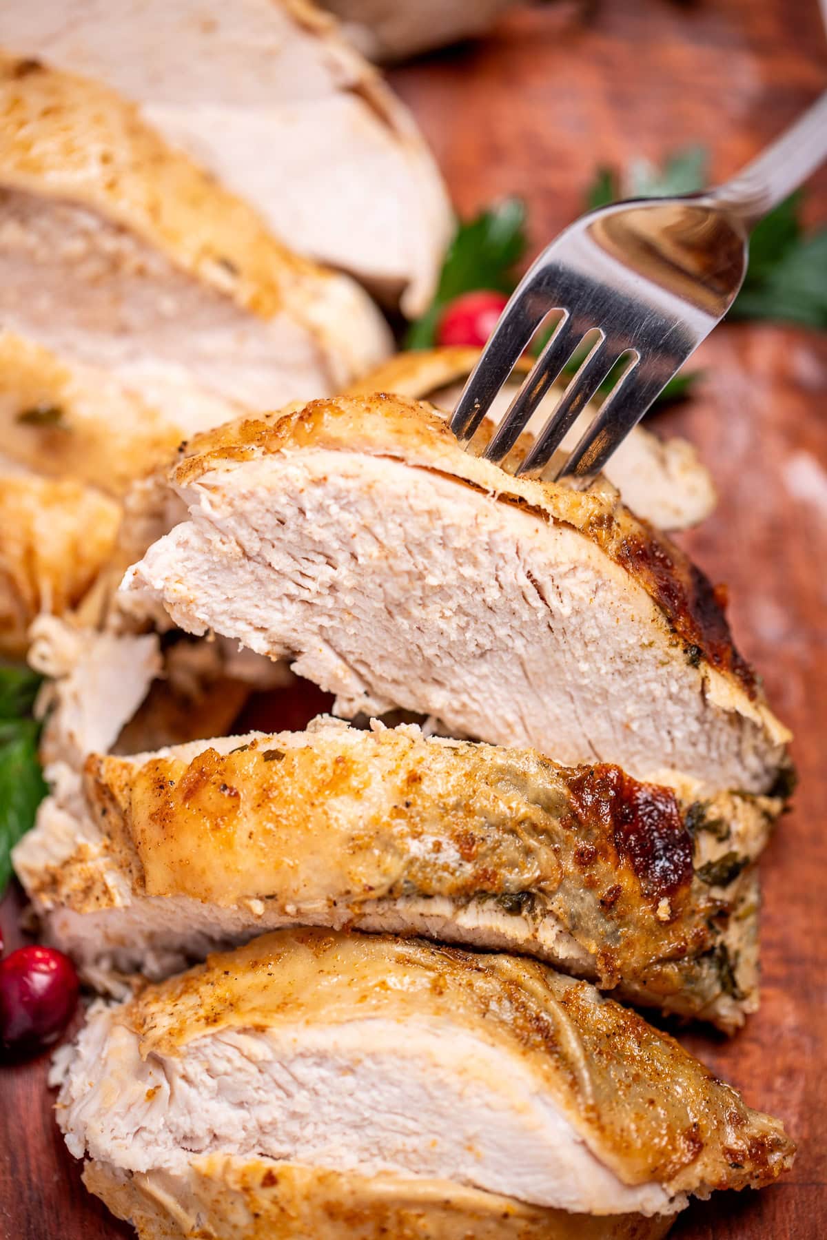 A cutting board with sliced turkey breast next to parsley and cranberries, with a fork picking a piece up.