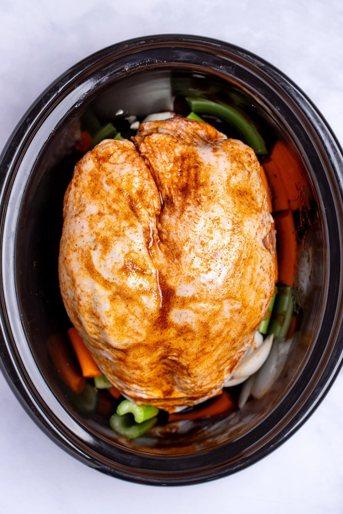 A slow cooker with a raw seasoned bone-in turkey breast resting on top of vegetables before being cooked.