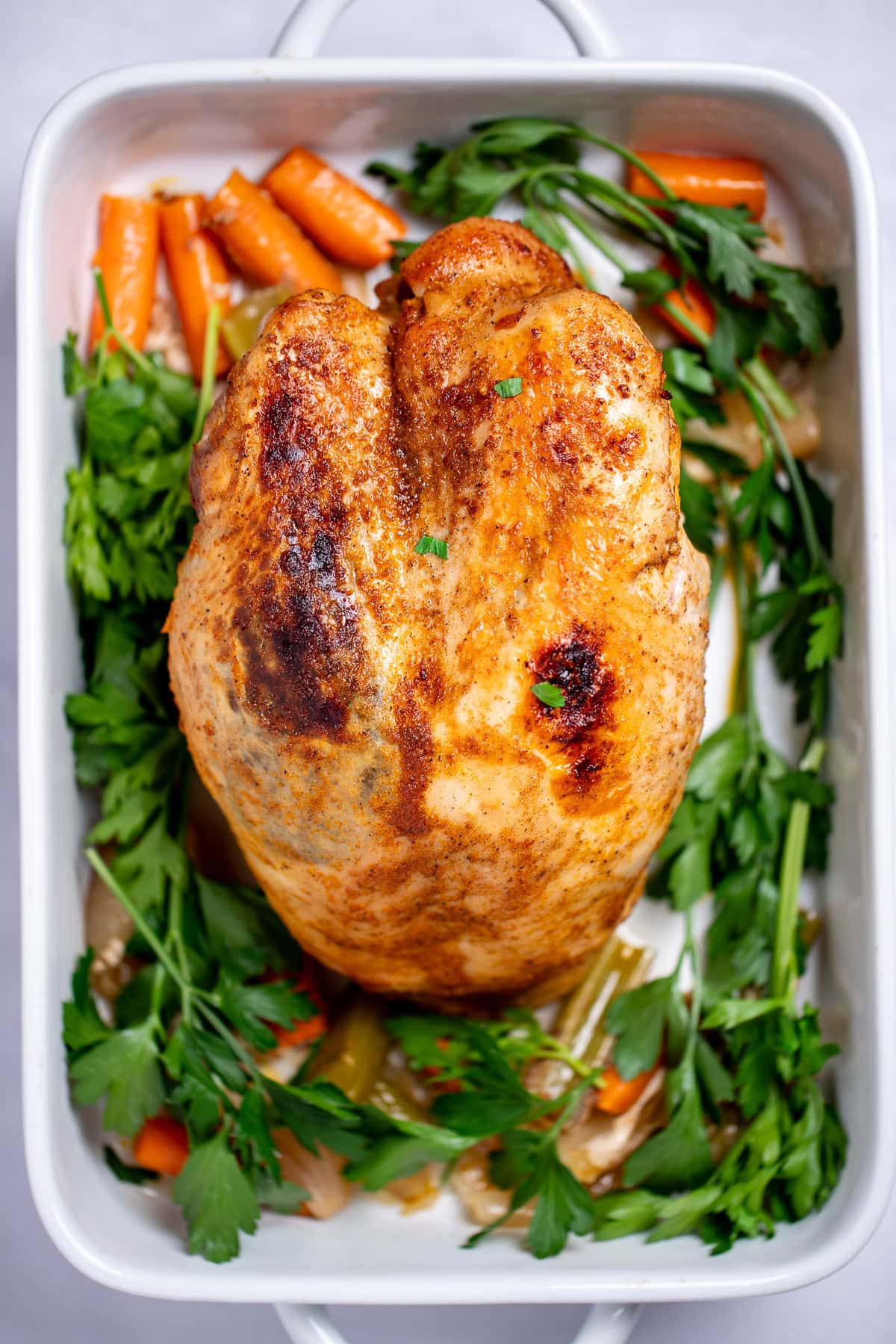 A baking dish with a cooked bone-in turkey breast surrounded by carrot, celery, onion, and parsley.