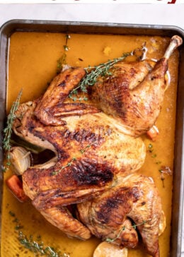 Pinterest pin with a roasting pan with a golden brown spatchcocked turkey resting on top of lemon and vegetables, with melted butter and fresh thyme on the bottom of the pan, and a kitchen towel on the table next to the roasting pan.