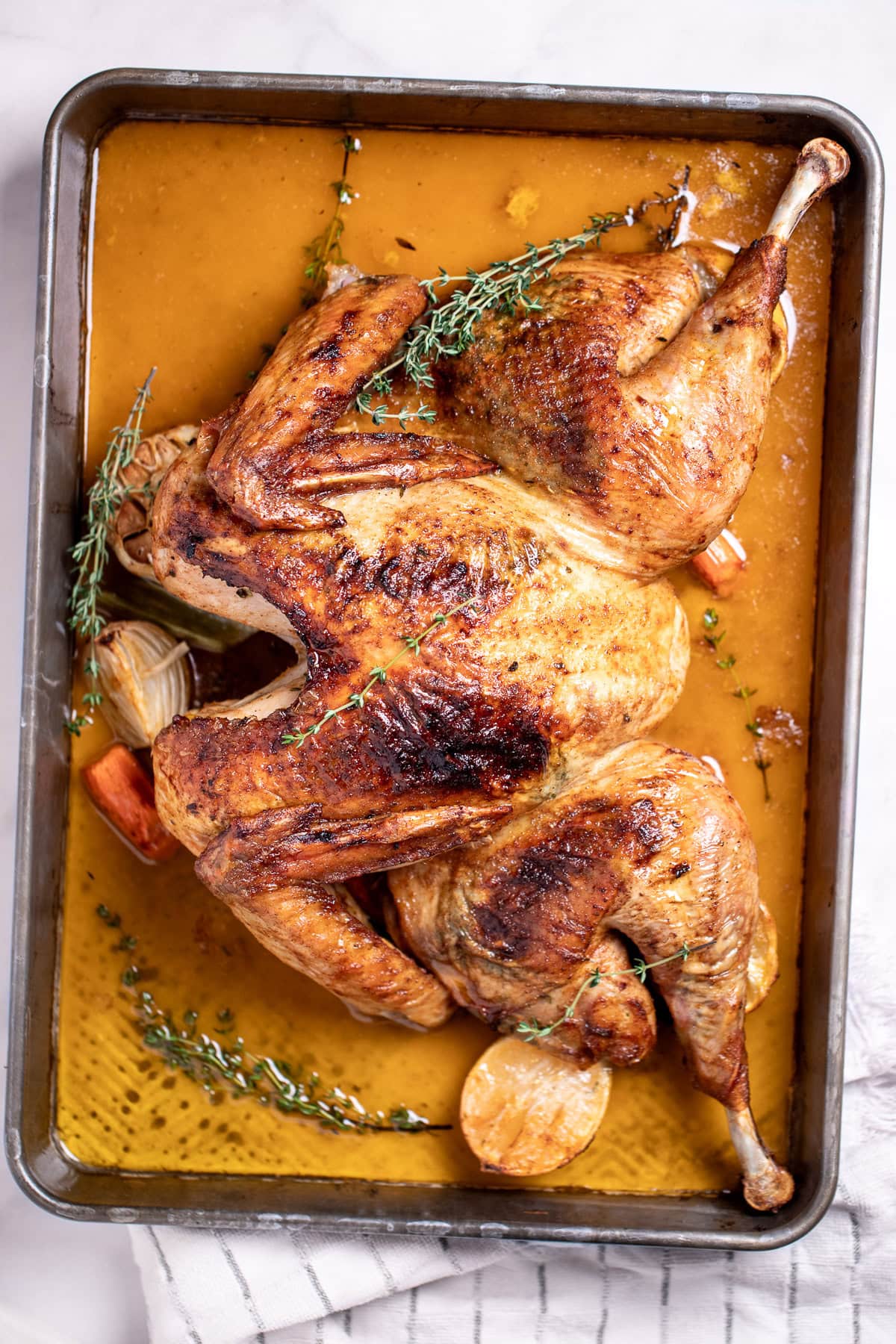 A roasting pan with a golden brown spatchcocked turkey resting on top of lemon and vegetables, with melted butter and fresh thyme on the bottom of the pan, and a kitchen towel on the table next to the roasting pan.