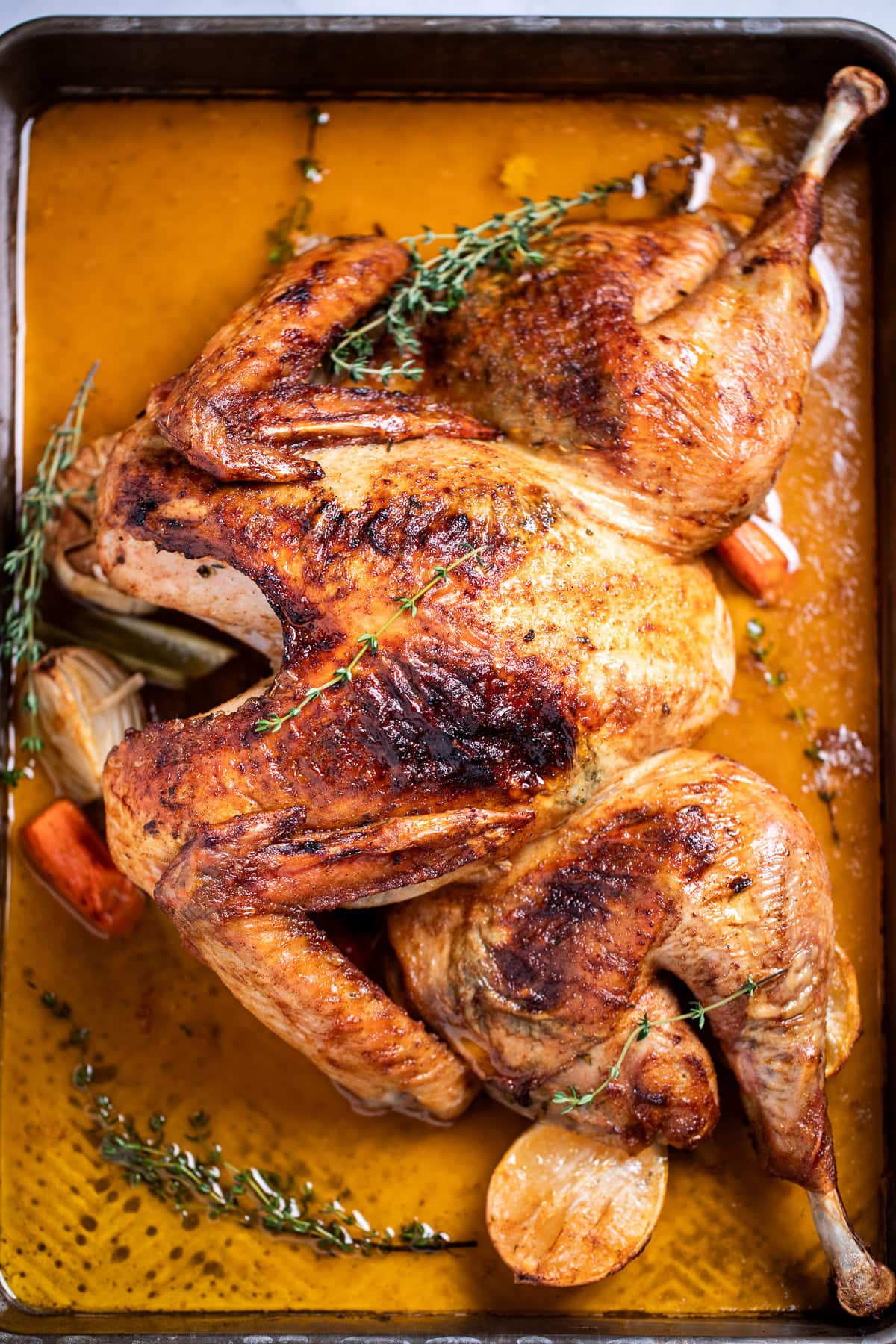 A roasting pan with a golden brown spatchcocked turkey resting on top of lemon and vegetables, with melted butter and fresh thyme on the bottom of the pan, and a kitchen towel on the table next to the roasting pan.
