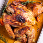 A roasting pan with a golden brown spatchcocked turkey resting on top of lemon and vegetables, with melted butter and fresh thyme on the bottom of the pan, and a kitchen towel on the table next to the roasting pan.