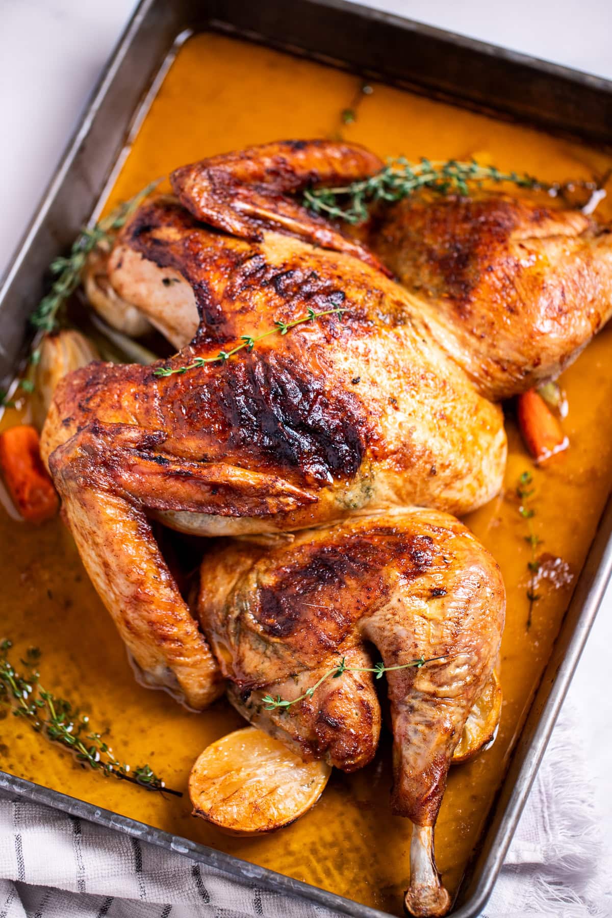 A roasting pan with a golden brown spatchcocked turkey resting on top of lemon and vegetables, with melted butter and fresh thyme on the bottom of the pan, and a kitchen towel on the table next to the roasting pan.