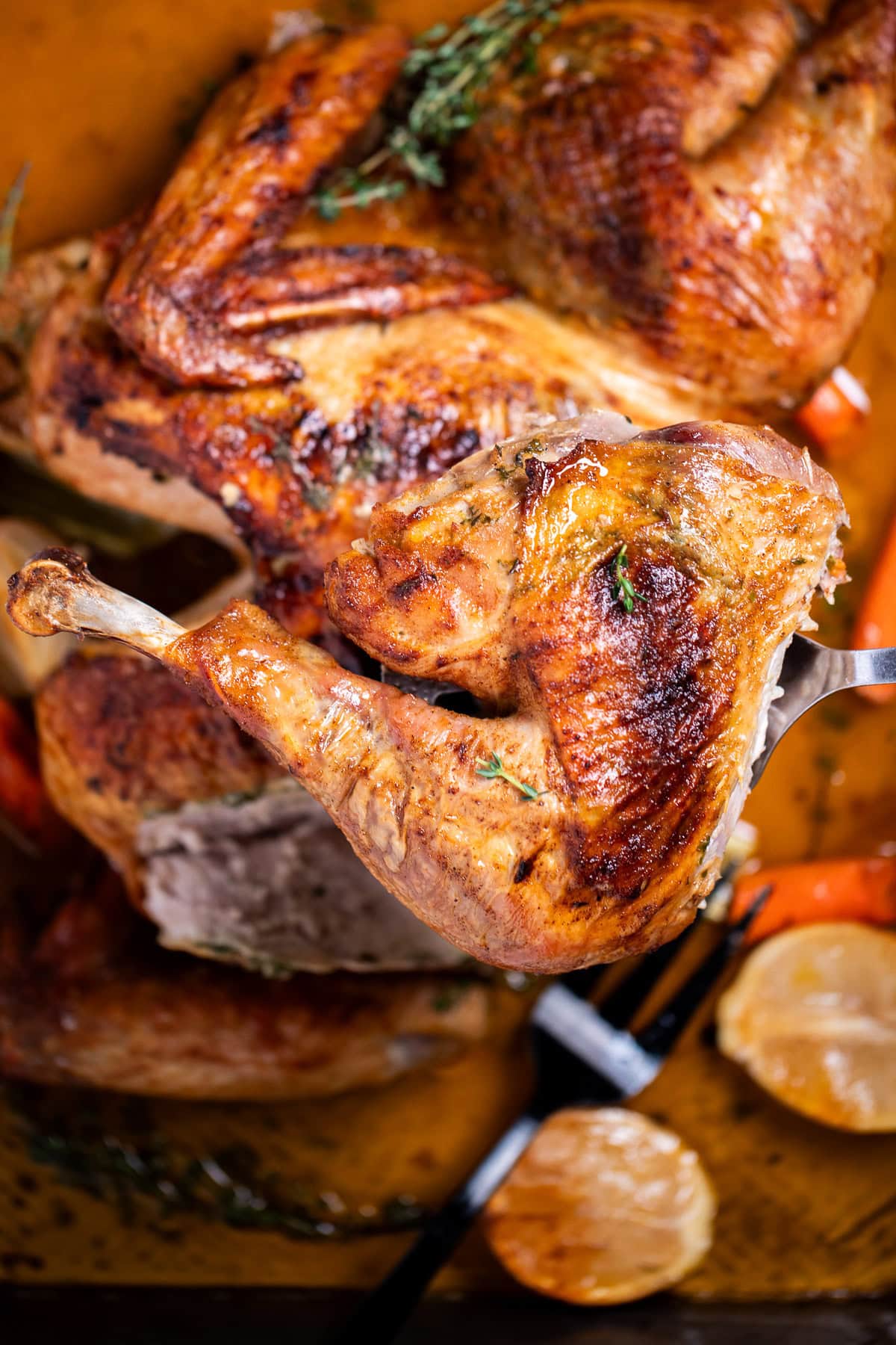 A roasting pan with a golden brown spatchcocked turkey resting on top of lemon and vegetables, with melted butter and fresh thyme on the bottom of the pan, and a turkey leg being lifted up by a spatula.