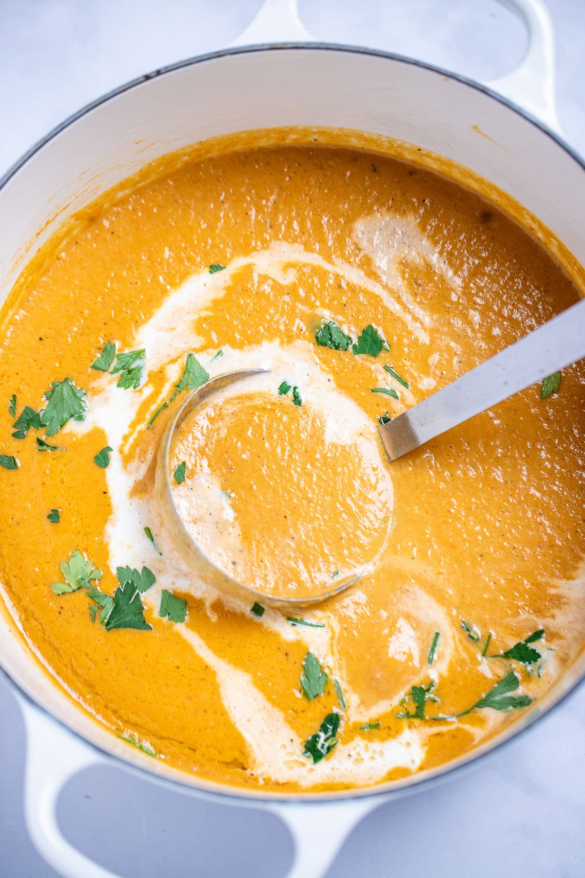 A dutch oven with creamy sweet potato soup topped with a drizzle of cream and chopped fresh cilantro, with a ladle resting in the soup, on a table.