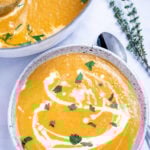 A bowl of creamy sweet potato soup topped with a drizzle of cream and chopped fresh cilantro, on a table next to a spoon and a sprig of fresh thyme, with a dutch oven of soup in the background.