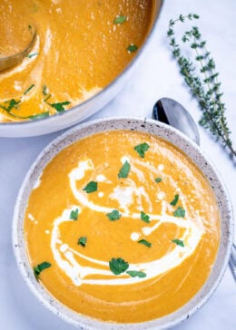 A bowl of creamy sweet potato soup topped with a drizzle of cream and chopped fresh cilantro, on a table next to a spoon and a sprig of fresh thyme, with a dutch oven of soup in the background.