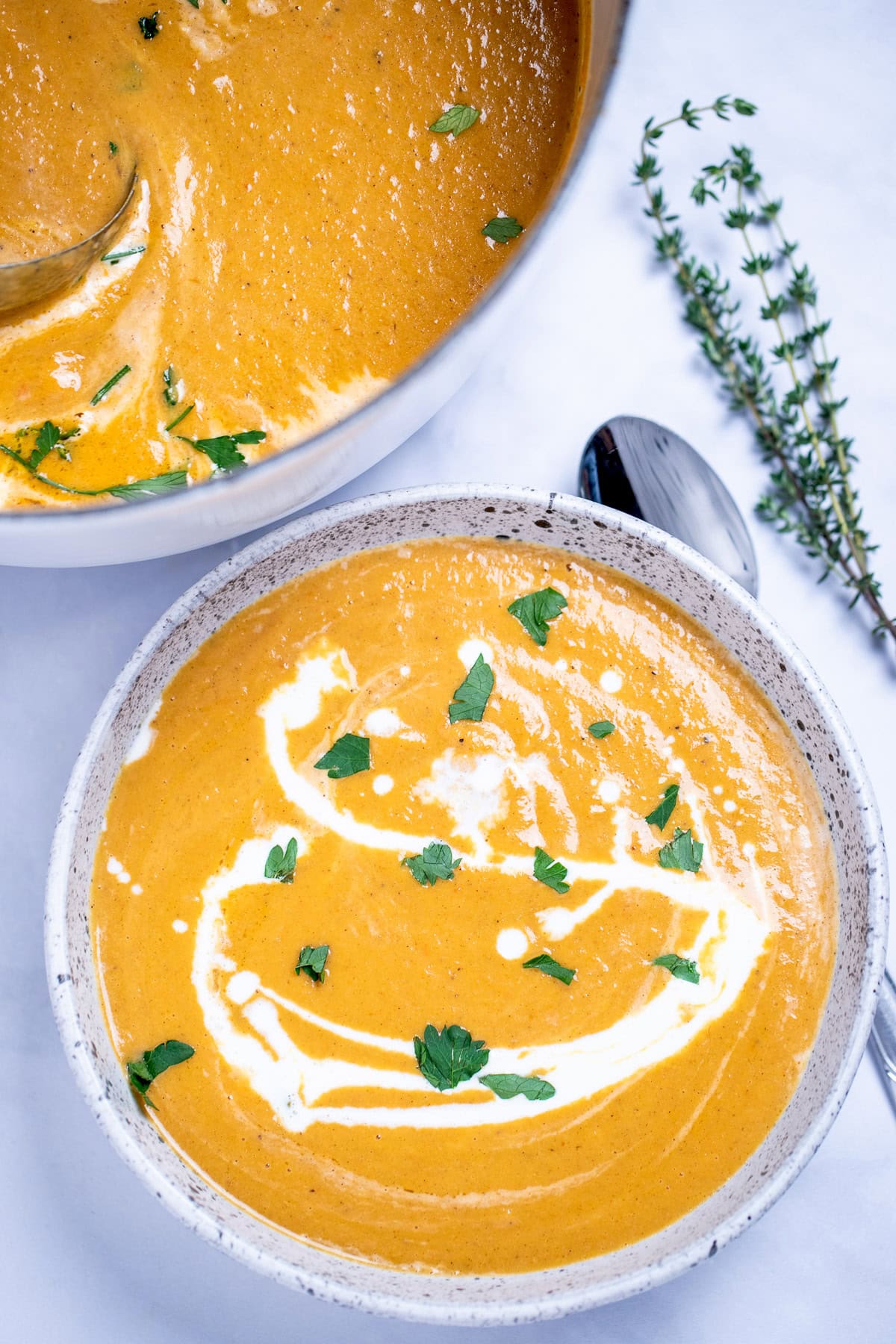A bowl of creamy sweet potato soup topped with a drizzle of cream and chopped fresh cilantro, on a table next to a spoon and a sprig of fresh thyme, with a dutch oven of soup in the background.