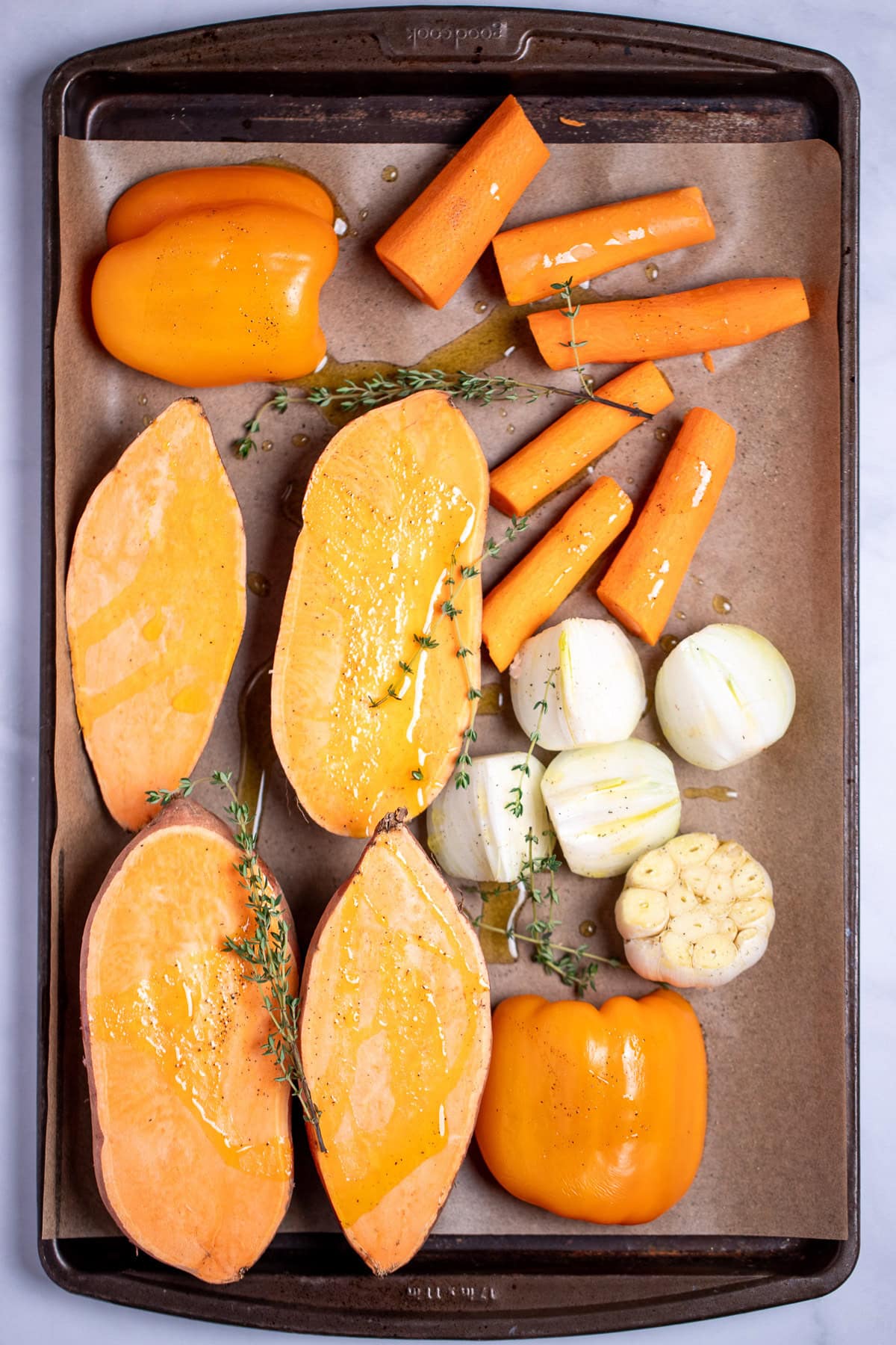 A sheet pan lined with parchment paper with two sweet potatoes cut in half vertically, two halves of a bell pepper, a quartered onion, chopped carrots, and a garlic bulb cut open, topped with fresh thyme and olive oil, before being roasted.