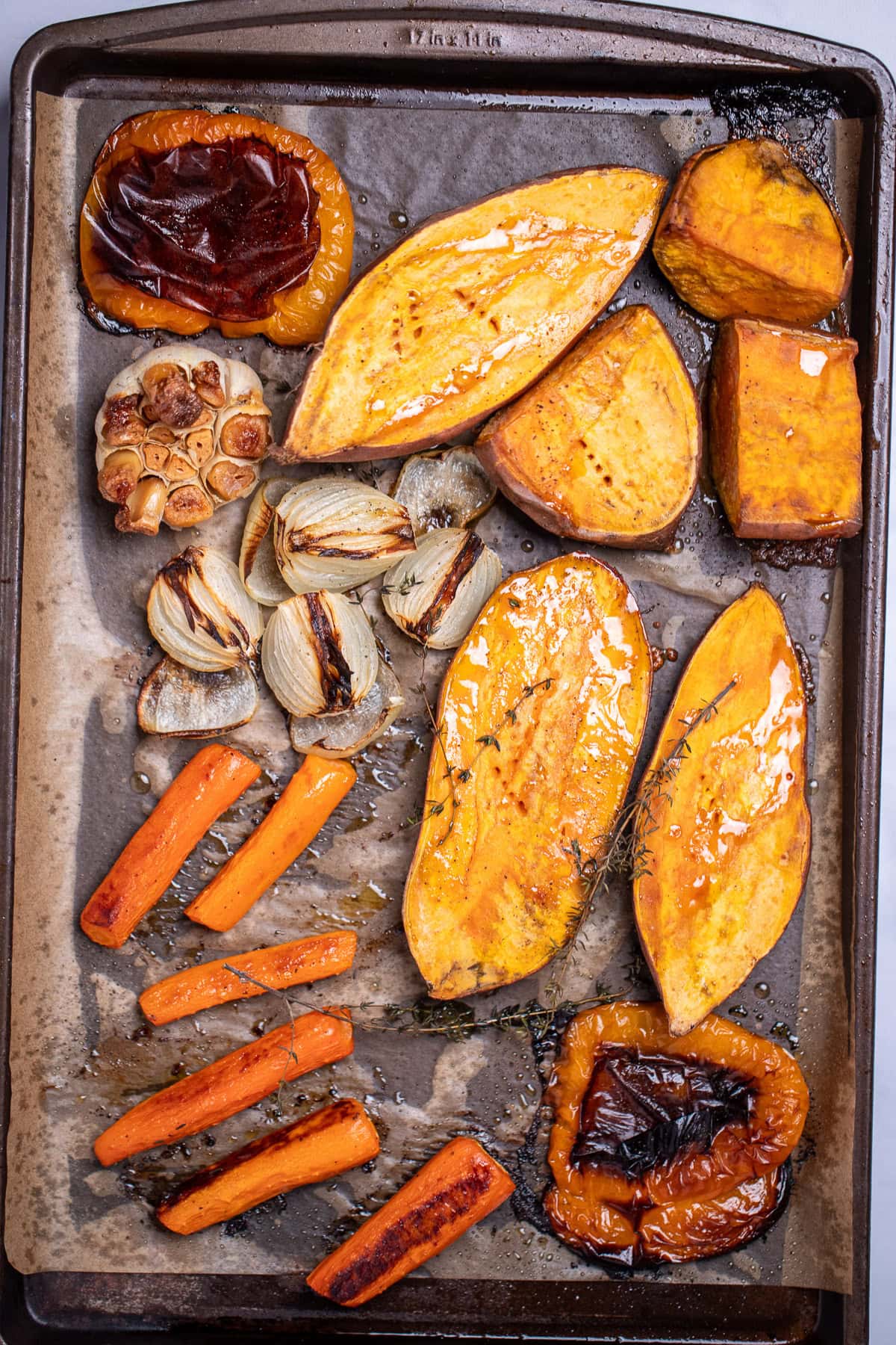 A sheet pan lined with parchment paper with two sweet potatoes cut in half vertically, two halves of a bell pepper, a quartered onion, chopped carrots, and a garlic bulb cut open, topped with fresh thyme and olive oil, after being roasted.