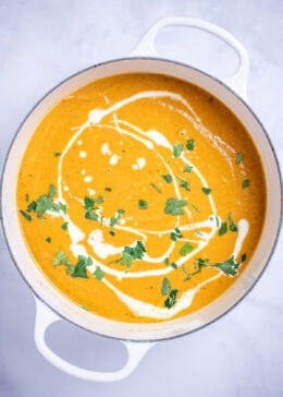 A dutch oven with creamy sweet potato soup topped with a drizzle of cream and chopped fresh cilantro, on a table.