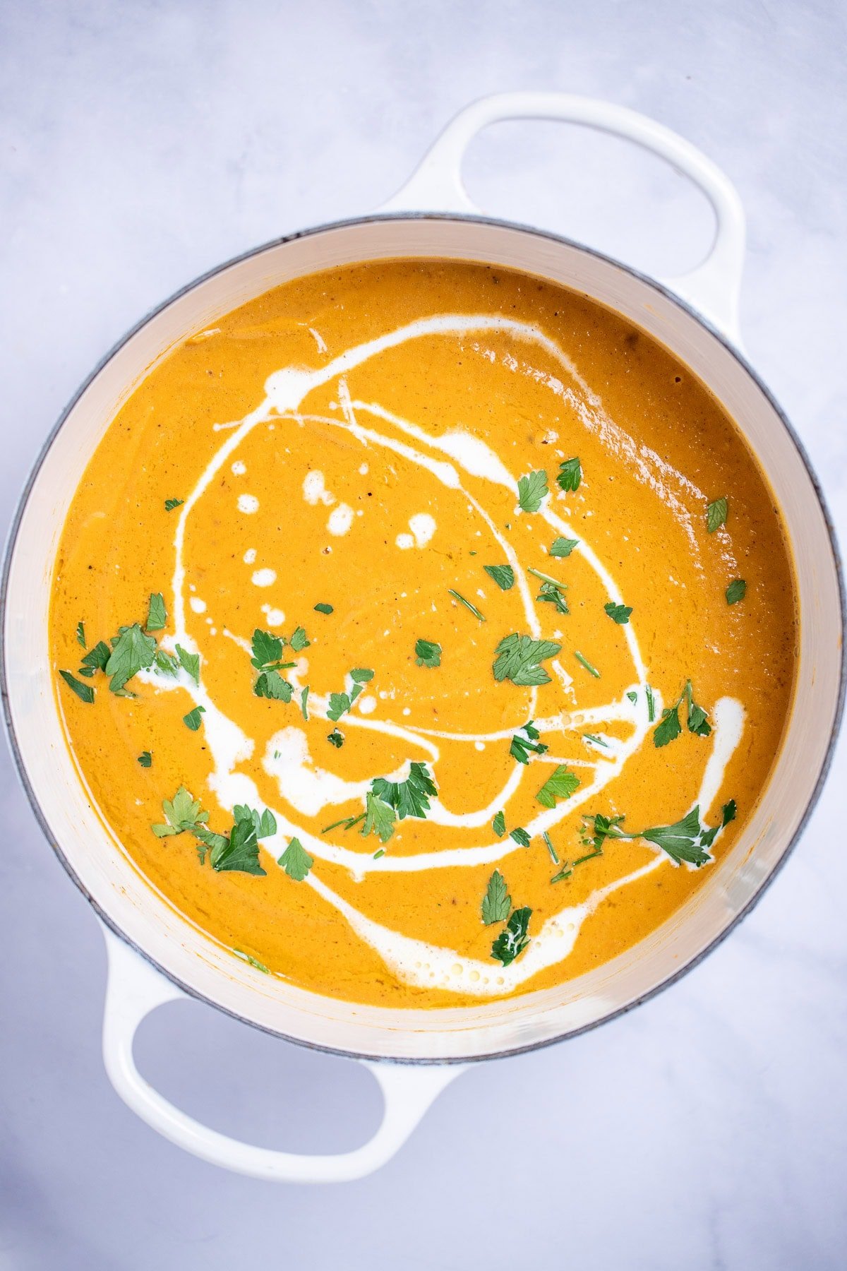 A dutch oven with creamy sweet potato soup topped with a drizzle of cream and chopped fresh cilantro, on a table.