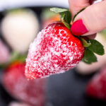 A hand holding a sugar coated champagne strawberry over a bowl of strawberries.