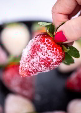 A hand holding a sugar coated champagne strawberry over a bowl of strawberries.