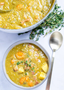 A bowl with split pea soup with ham, carrots, and potatoes, with thyme on top, next to a spoon and in front of more fresh thyme and a dutch oven.