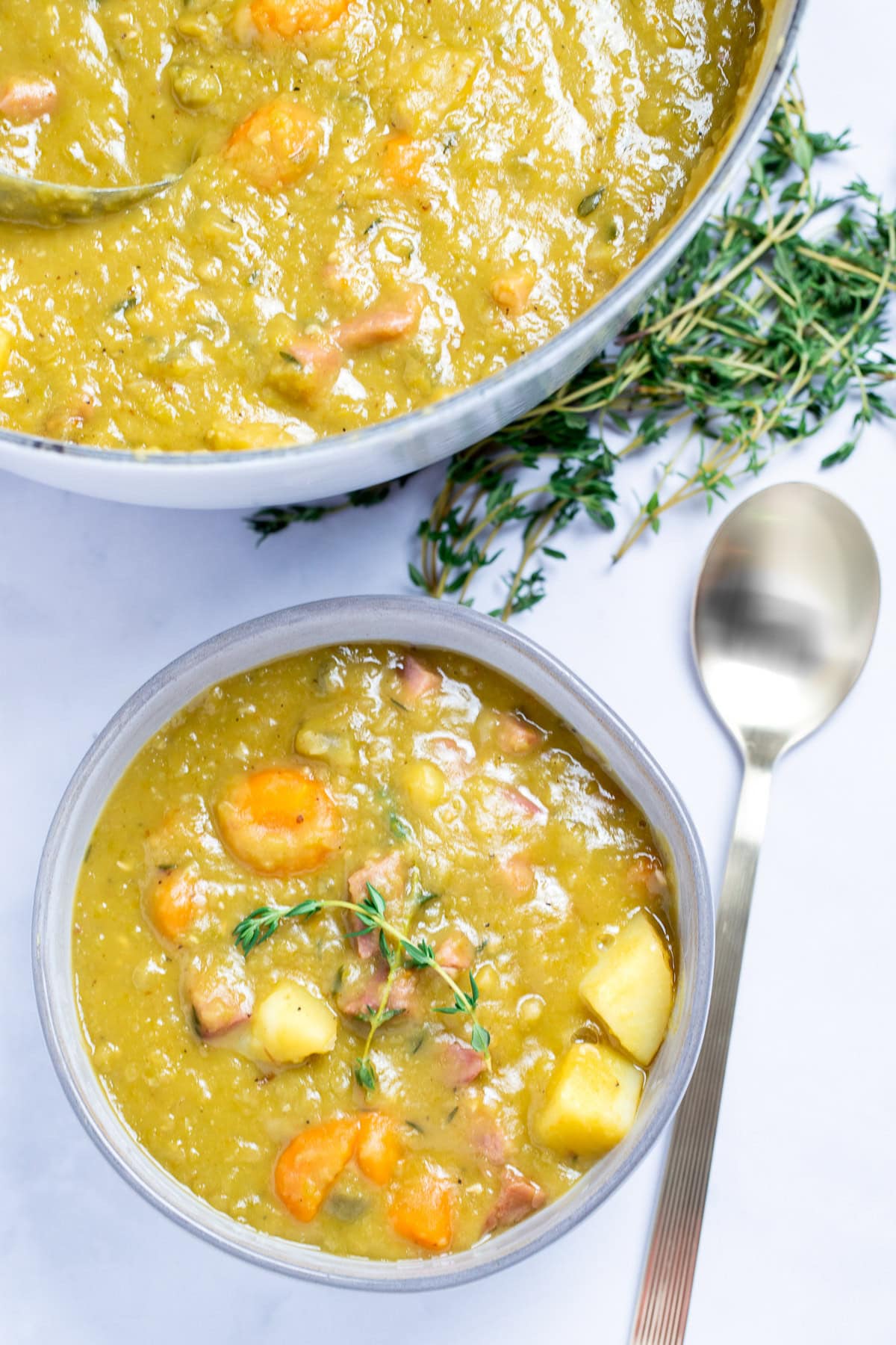 A bowl with split pea soup with ham, carrots, and potatoes, with thyme on top, next to a spoon and in front of more fresh thyme and a dutch oven.