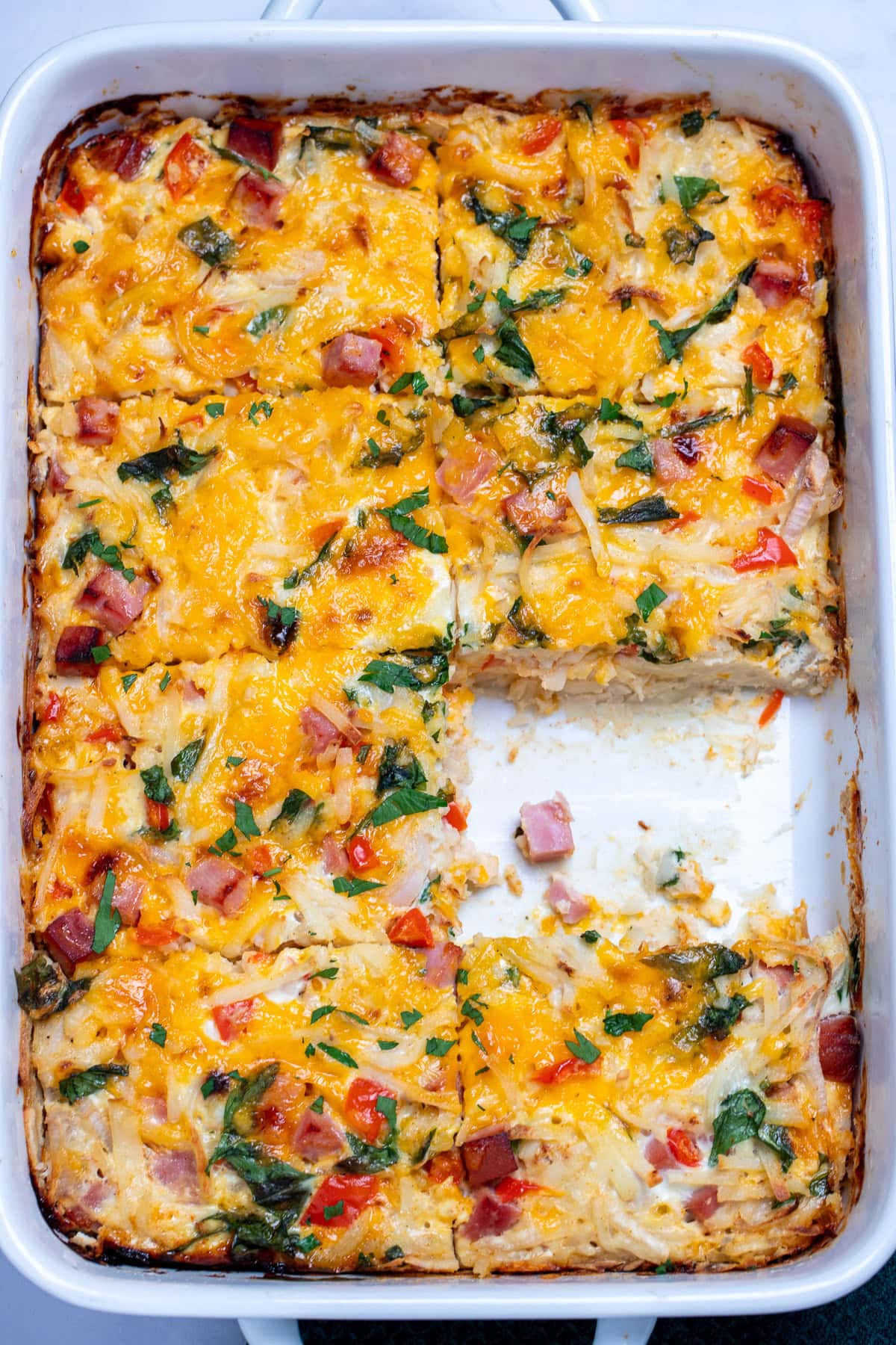A casserole dish with hashbrown breakfast casserole topped with melted cheese and fresh parsley, with a piece missing.