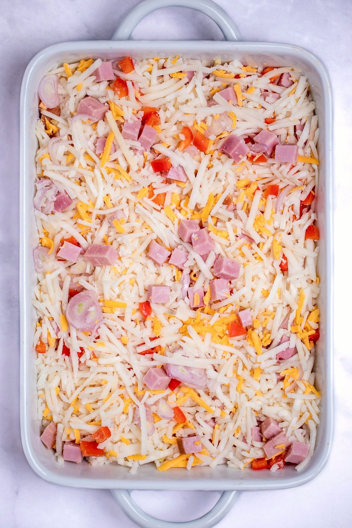 A casserole dish with hashbrowns, cheese, peppers and ham mixed together before cooking.