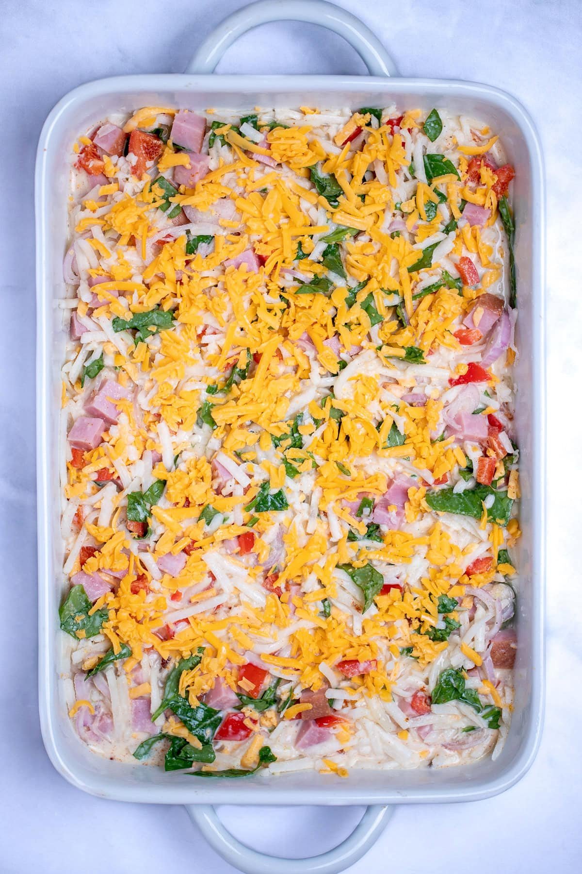 A casserole dish with hashbrowns, cheese, peppers, spinach, eggs, and ham mixed together before cooking.