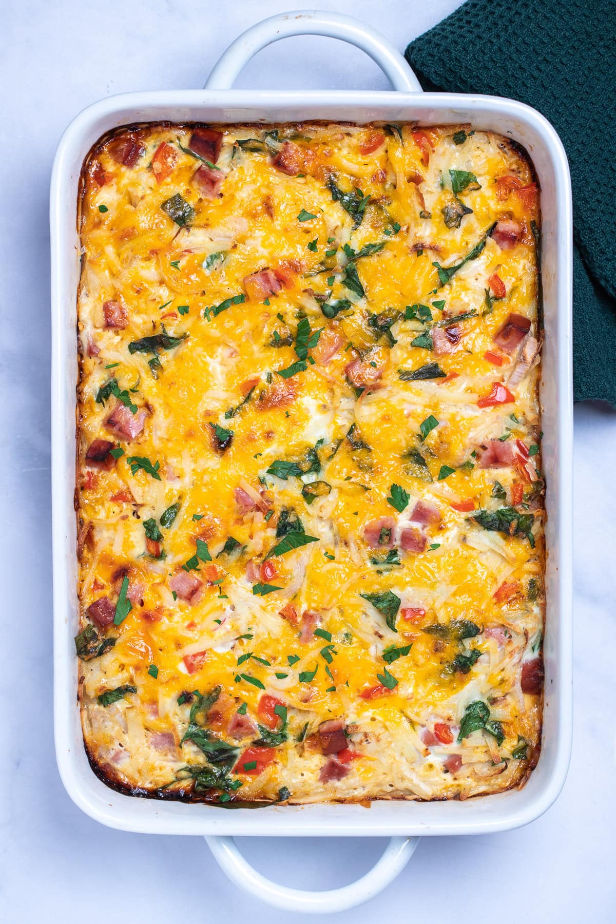 A casserole dish with hashbrown breakfast casserole topped with melted cheese and fresh parsley, next to a kitchen towel.