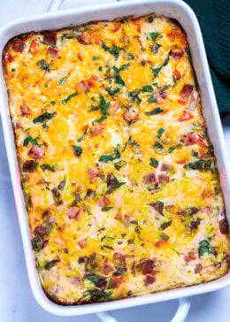 A casserole dish with hashbrown breakfast casserole topped with melted cheese and fresh parsley, next to a kitchen towel.
