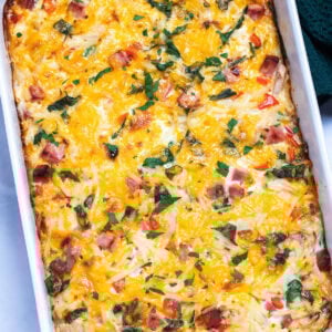 A casserole dish with hashbrown breakfast casserole topped with melted cheese and fresh parsley, next to a kitchen towel.