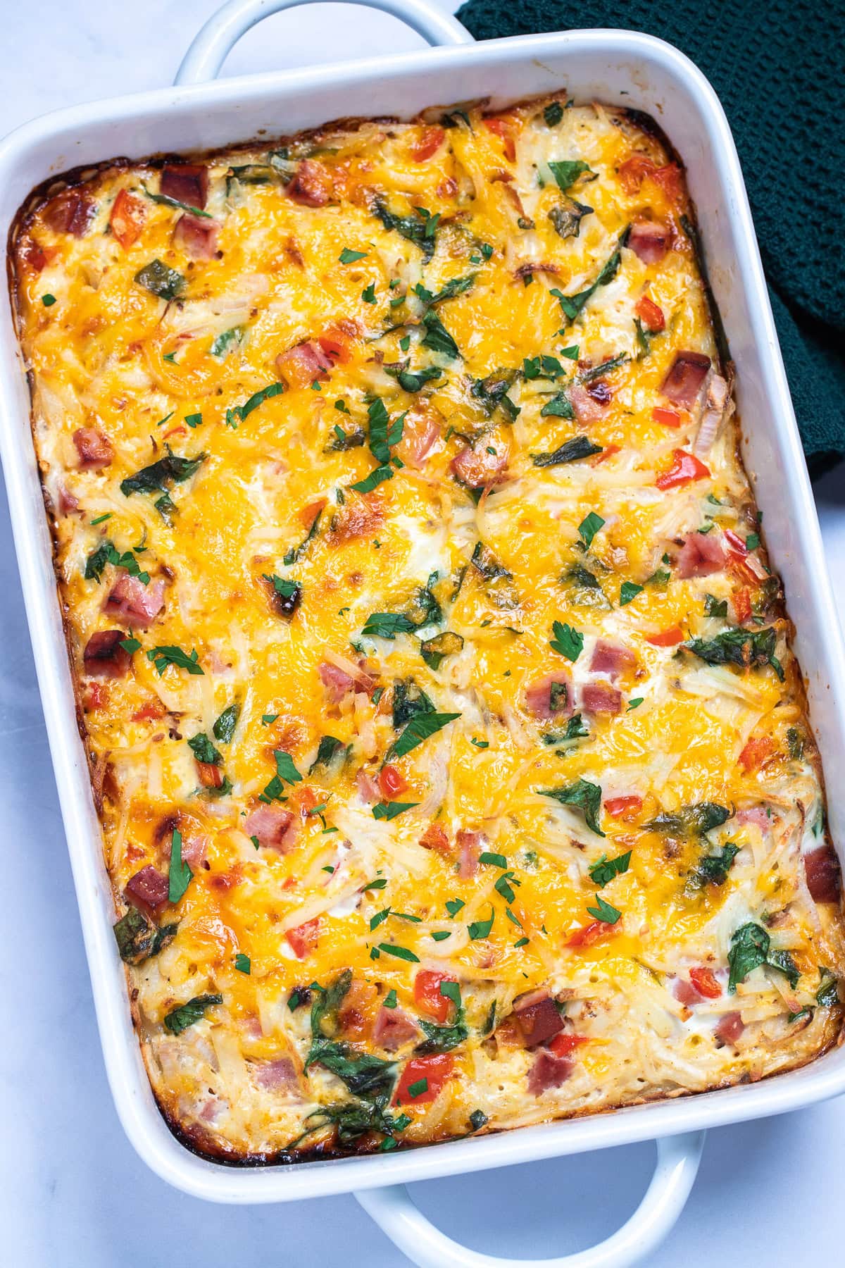 A casserole dish with hashbrown breakfast casserole topped with melted cheese and fresh parsley, next to a kitchen towel.