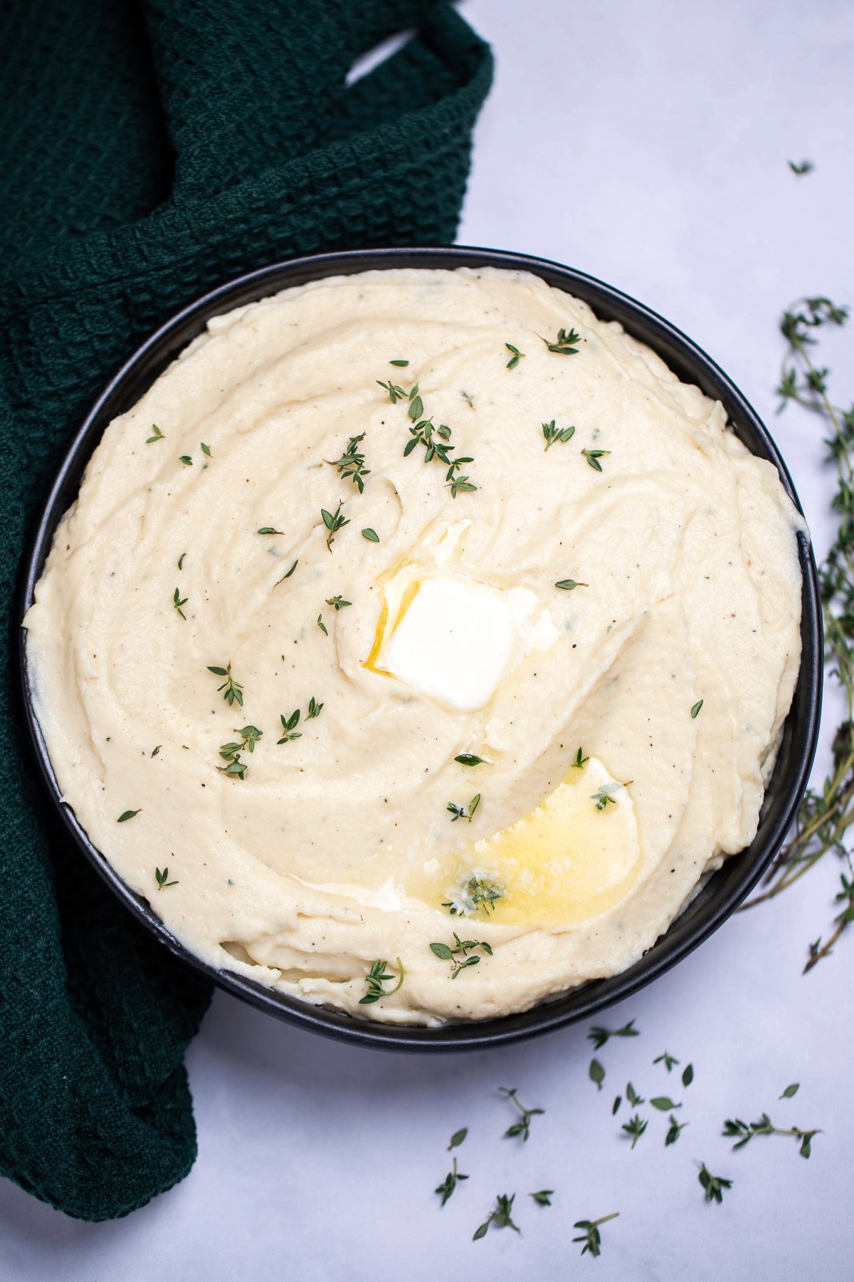 A bowl of parsnip puree topped with butter and thyme, next to a towel on a table.