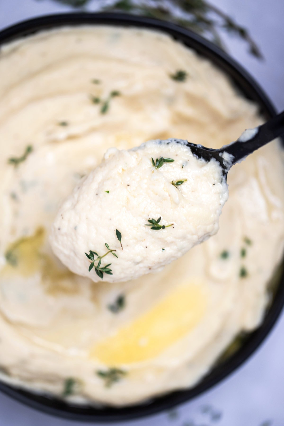 A bowl of parsnip puree topped with butter and thyme, next to fresh thyme on a table, and a spoon lifting some up.