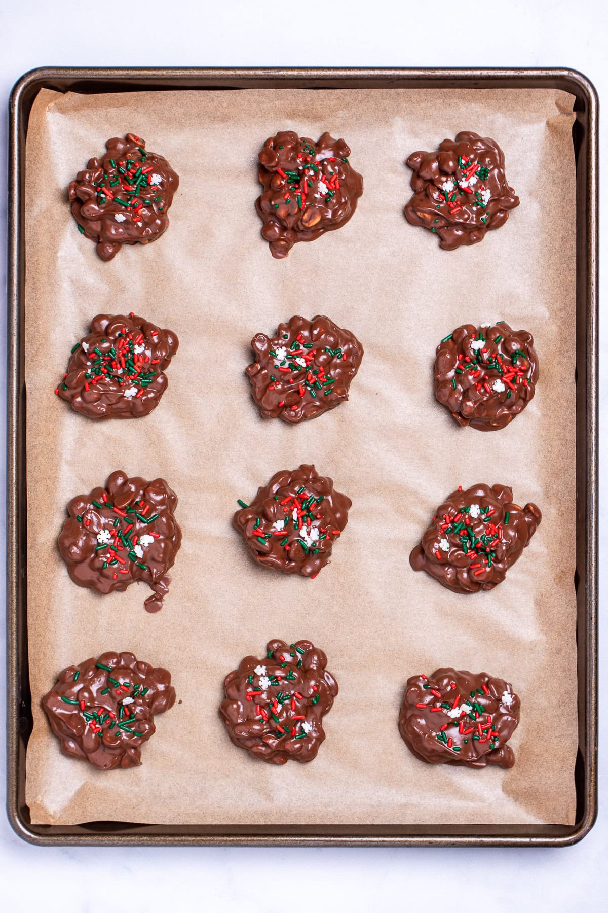 A sheet pan lined with parchment paper with chocolate peanut clusters topped with sprinkles before they set.