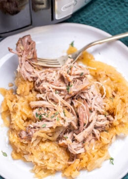 A plate with shredded pork shoulder over sauerkraut with a fork resting on a plate, in front of a slow cooker.