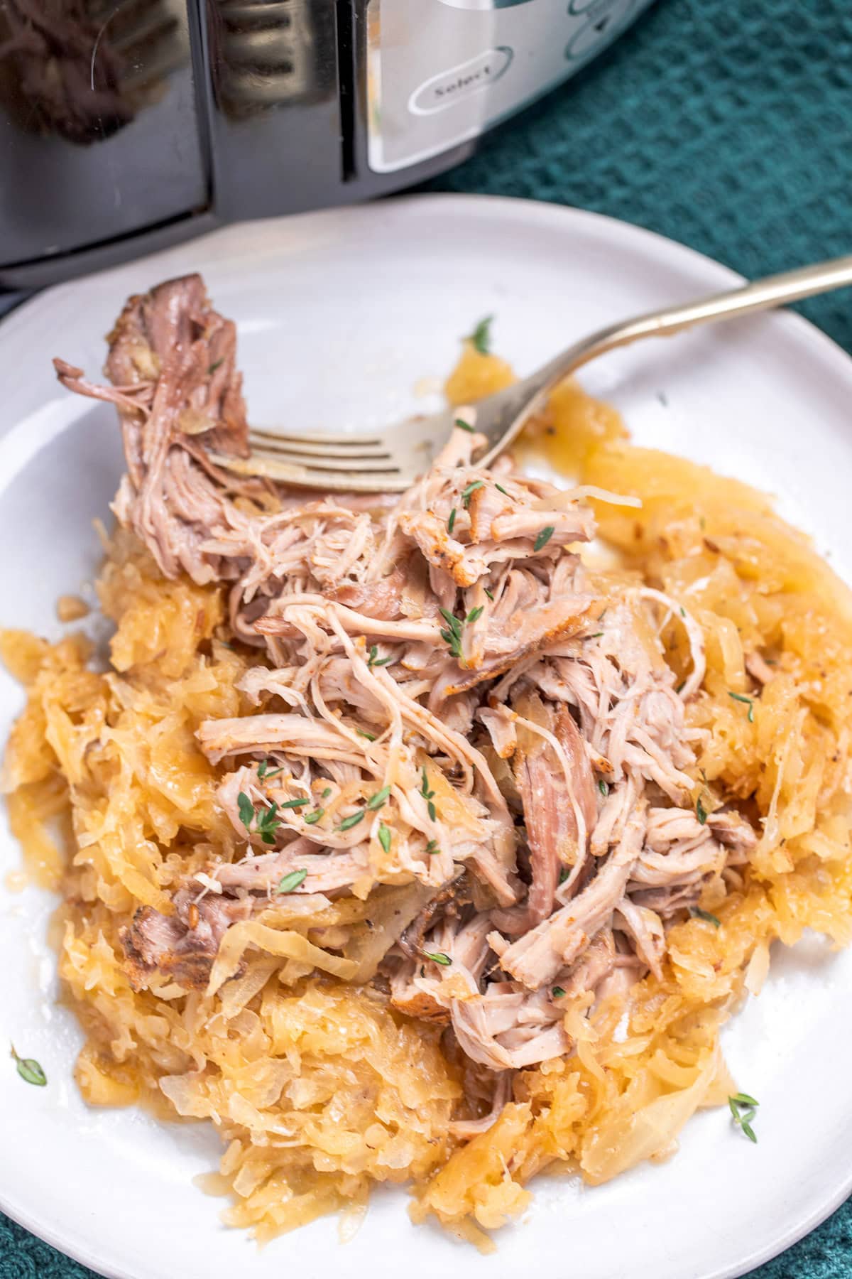 A plate with shredded pork shoulder over sauerkraut with a fork resting on a plate, in front of a slow cooker.