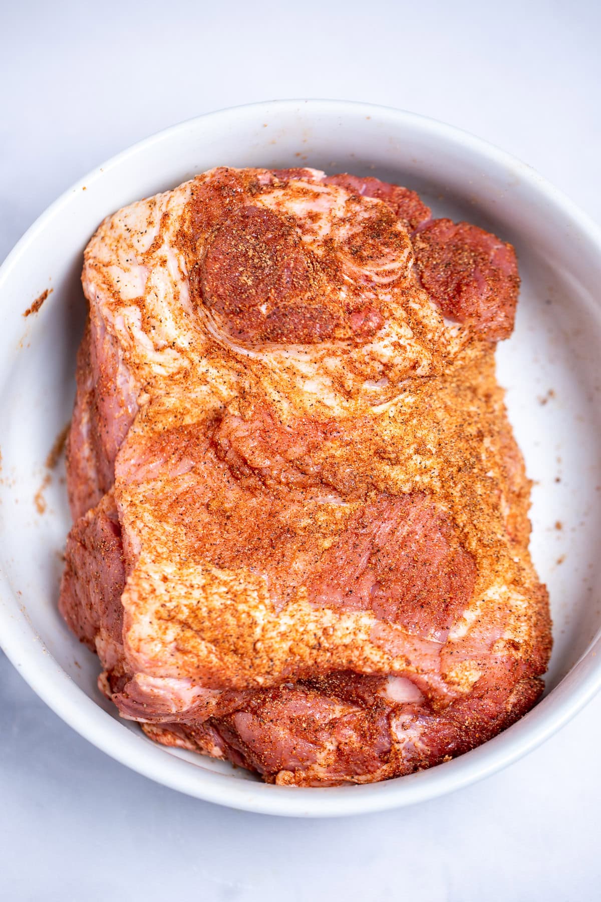 A bowl with a raw bone-in pork shoulder with seasoning rub all over it.