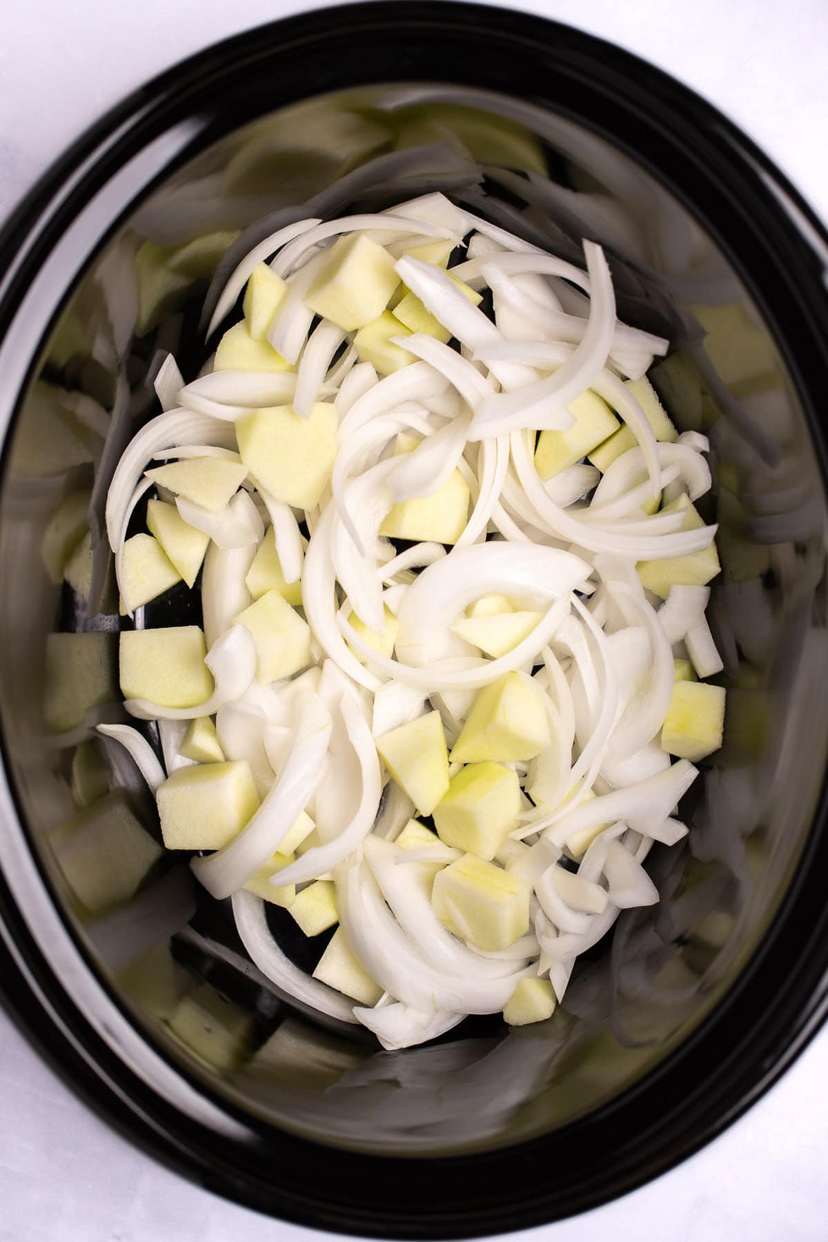 A slow cooker with onion and apple pieces on the bottom of it before being cooked.