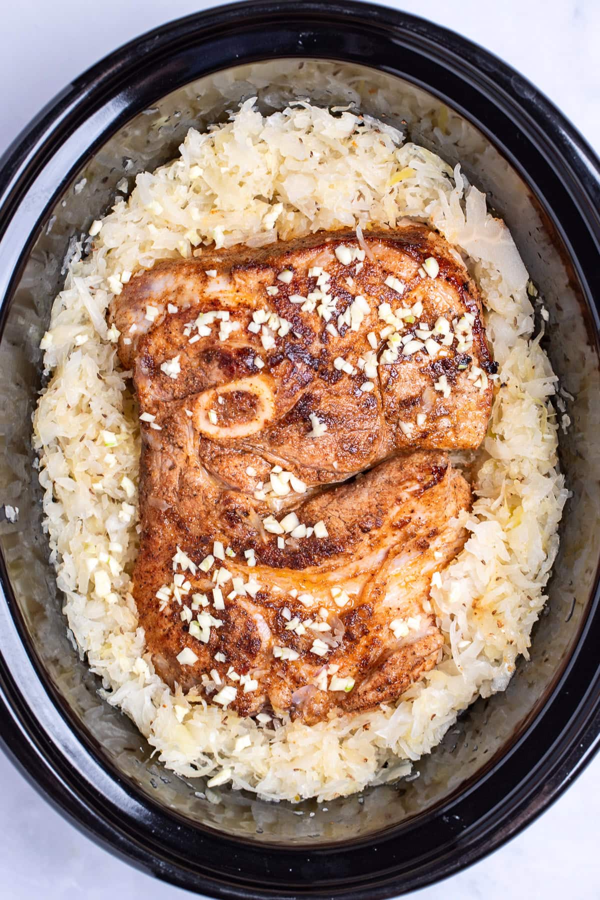 A slow cooker with a seared bone-in pork shoulder surrounded by sauerkraut.