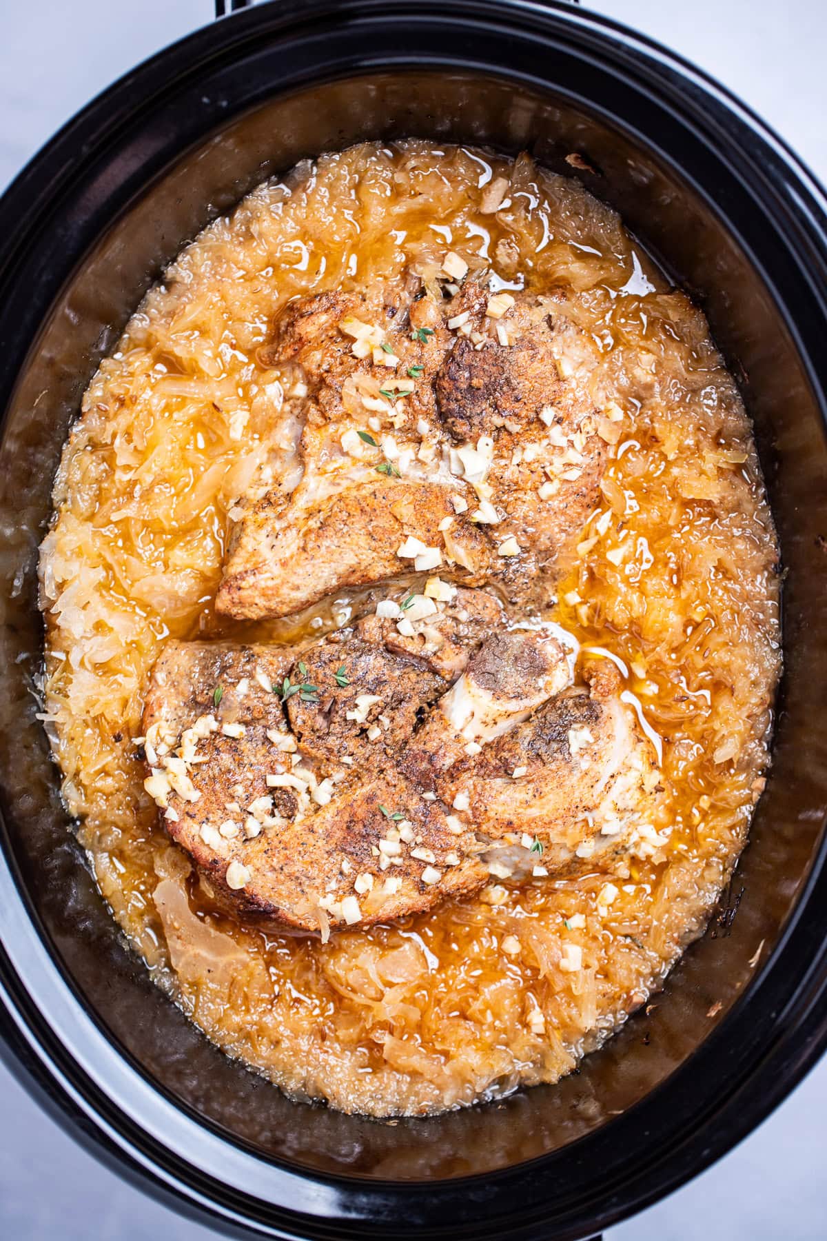 A slow cooker with cooked bone-in pork shoulder surrounded by sauerkraut and juices.