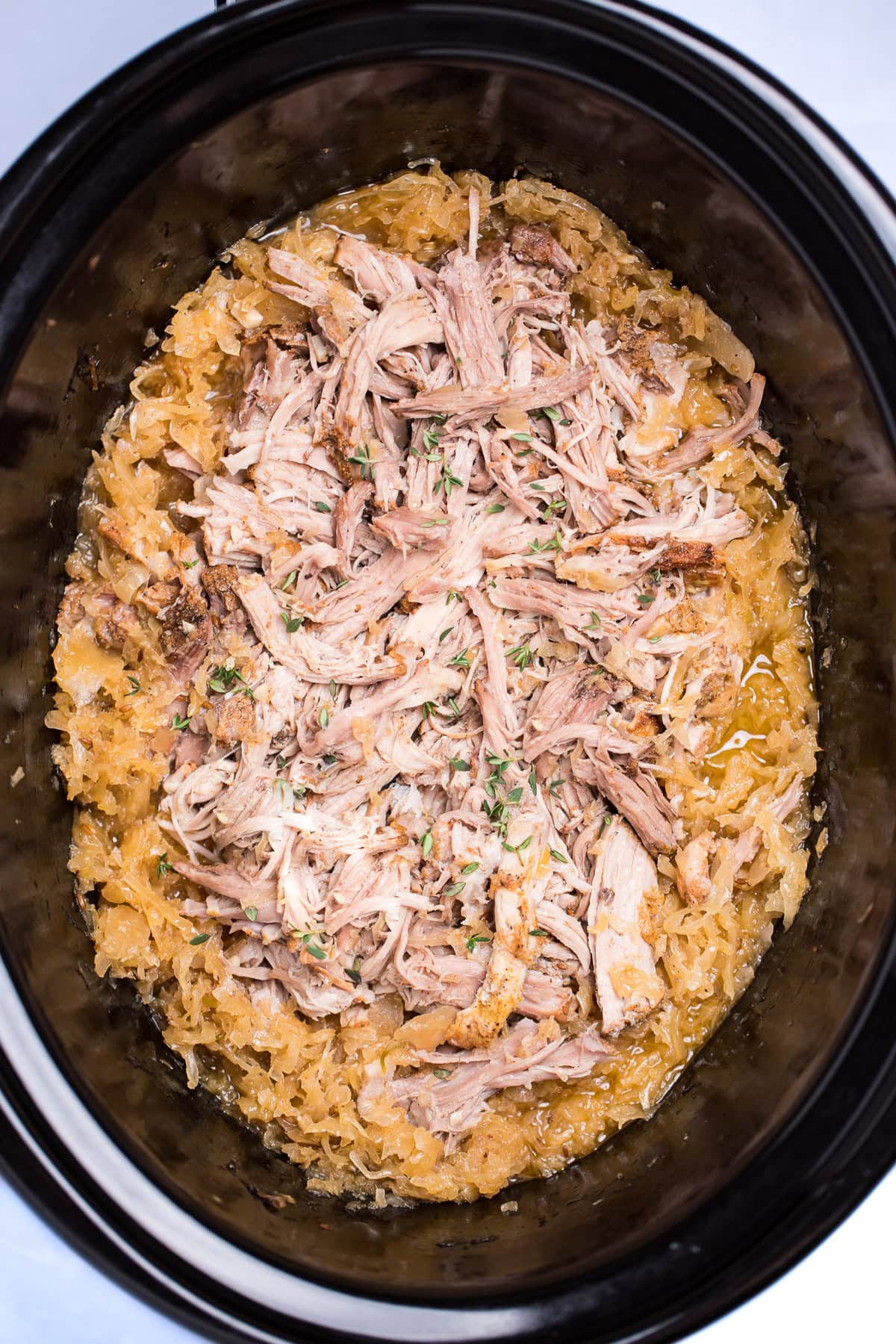 A slow cooker with cooked shredded bone-in pork shoulder surrounded by sauerkraut and juices.