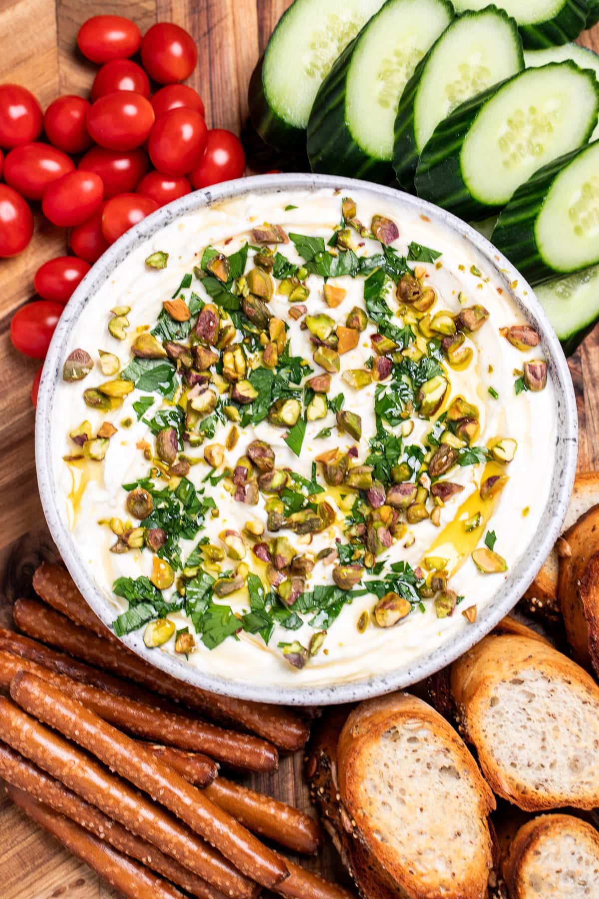 A bowl of whipped feta dip topped with chopped parsley, pistachios, olive oil, and honey on a wooden cutting board, surrounded by gluten free pretzels, gluten free toasted baguette slices, sliced cucumbers, and cherry tomatoes.