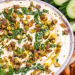 A bowl of whipped feta dip topped with chopped parsley, pistachios, olive oil, and honey on a wooden cutting board, surrounded by gluten free pretzels, gluten free toasted baguette slices, sliced cucumbers, and cherry tomatoes.