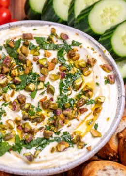 A bowl of whipped feta dip topped with chopped parsley, pistachios, olive oil, and honey on a wooden cutting board, surrounded by gluten free pretzels, gluten free toasted baguette slices, sliced cucumbers, and cherry tomatoes.