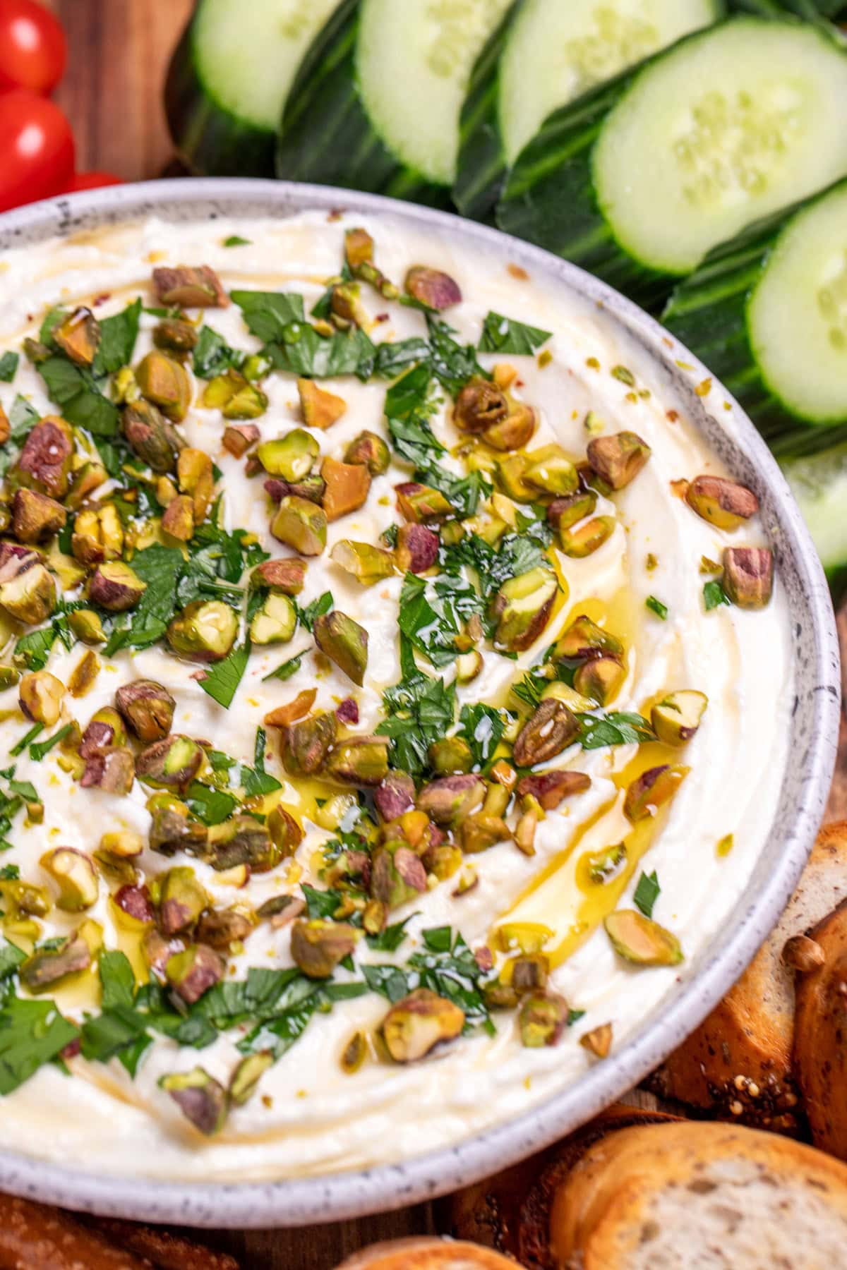 A bowl of whipped feta dip topped with chopped parsley, pistachios, olive oil, and honey on a wooden cutting board, surrounded by gluten free pretzels, gluten free toasted baguette slices, sliced cucumbers, and cherry tomatoes.