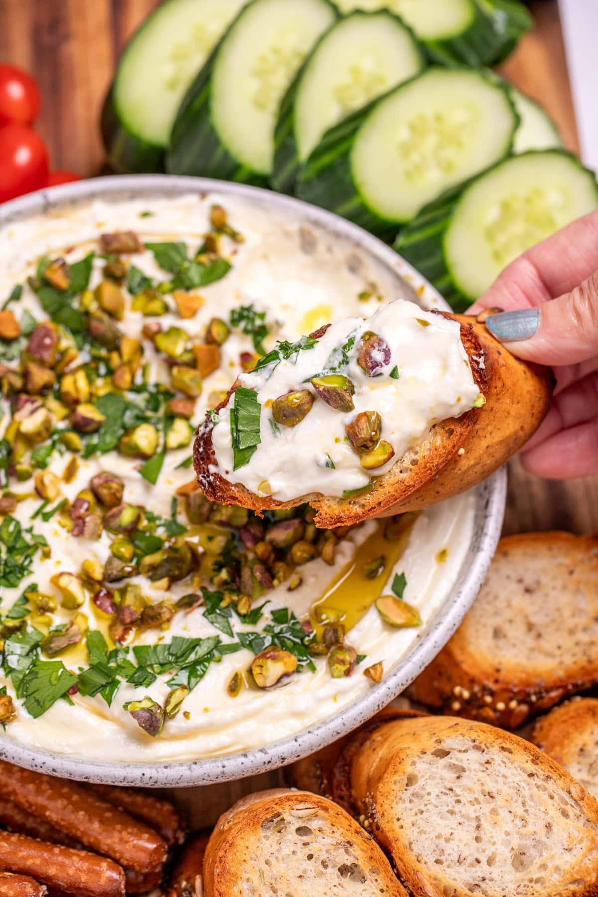 A bowl of whipped feta dip topped with chopped parsley, pistachios, olive oil, and honey on a wooden cutting board, surrounded by gluten free pretzels, gluten free toasted baguette slices, sliced cucumbers, and cherry tomatoes. A hand is holding a baguette slice covered in dip over the bowl.