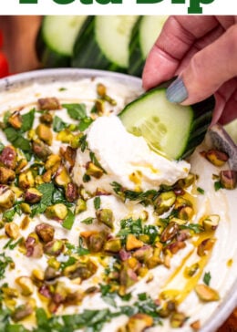 Pinterest pin with a bowl of whipped feta dip topped with chopped parsley, pistachios, olive oil, and honey on a wooden cutting board. A hand is holding a cucumber slice covered in dip over the bowl.