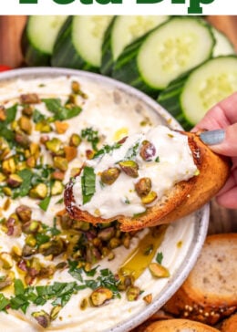 Pinterest pin with a bowl of whipped feta dip topped with chopped parsley, pistachios, olive oil, and honey on a wooden cutting board, surrounded by gluten free pretzels, gluten free toasted baguette slices, sliced cucumbers, and cherry tomatoes. A hand is holding a baguette slice covered in dip over the bowl.