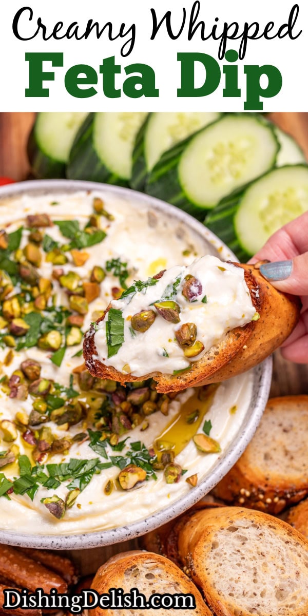 Pinterest pin with a bowl of whipped feta dip topped with chopped parsley, pistachios, olive oil, and honey on a wooden cutting board, surrounded by gluten free pretzels, gluten free toasted baguette slices, sliced cucumbers, and cherry tomatoes. A hand is holding a baguette slice covered in dip over the bowl.