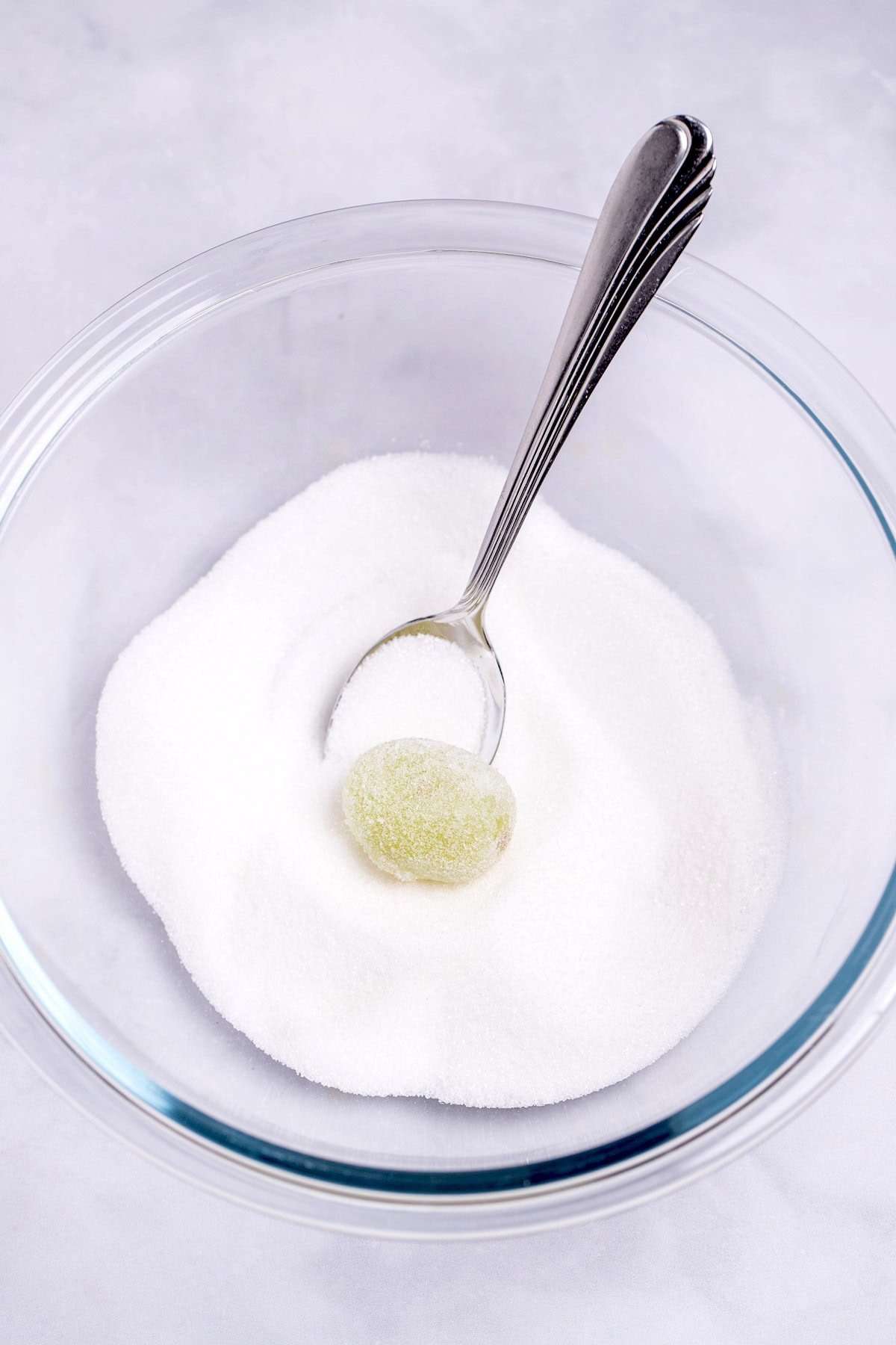 A glass bowl with sugar, and a spoon rolling a grape in the sugar.