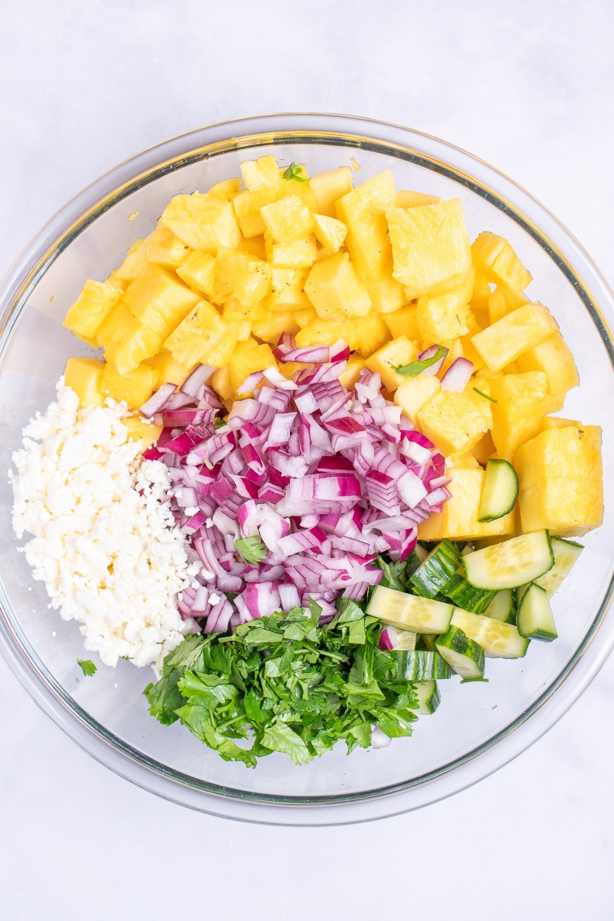 A glass bowl with chopped cucumber, pineapple, red onion, cilantro, and feta cheese.