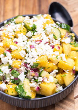 A bowl of pineapple cucumber salad topped with feta cheese sitting on a wooden cutting board with a spoon behind it.