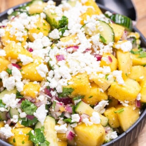 A bowl of pineapple cucumber salad topped with feta cheese sitting on a wooden cutting board with a spoon behind it.